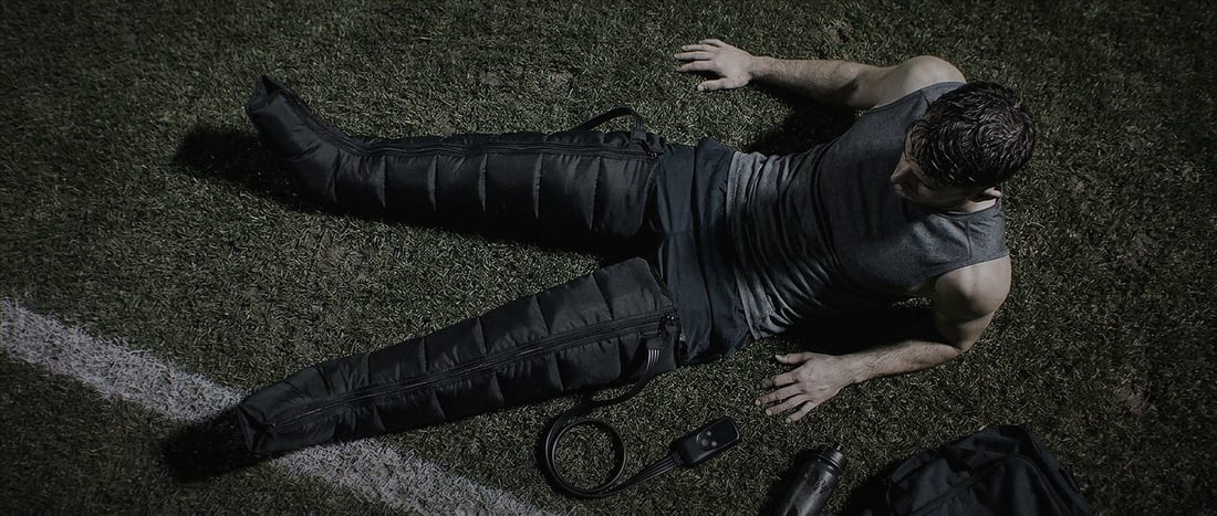 Seen from above, an athlete lies on a floodlit pitch wearing full-leg compression boots after a session.